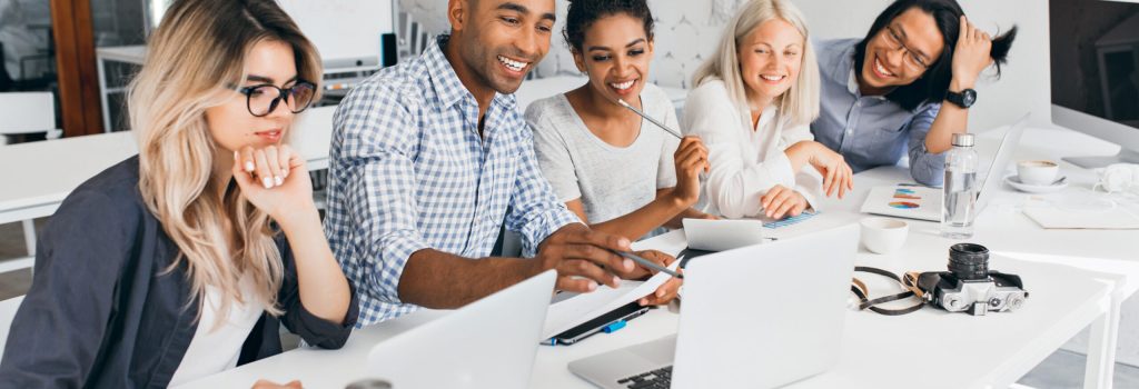 Smiling african student pointing with pencil at laptop screen. Concentrated blonde girl in glasses propping chin with hand while working with computer in office.